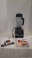 Front view of Vitamix 6300 blender with 64 oz blending container on top, power cord laid out. Two manuals/booklets placed to the left on table.