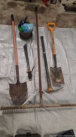 Photo showing five yard tools laid out on a white sheet including shovels, rake, and hand tools, plus a small pot with small bags inside