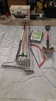 Photo showing six yard tools including rakes, hoe, bow saw, shovel, and packet of deer repellent powder on a tarp.