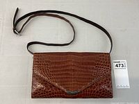 Full front view of brown crocodile leather envelope purse with long adjustable strap laid flat