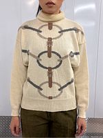 Front view of cream-colored turtleneck sweater showing bridle pattern in grey, taupe, and brown.