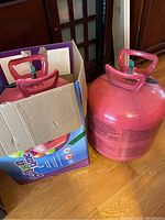 Close-up of one large pink Balloon Time helium tank partially inside its colorful branded box on a wooden floor.