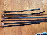 Six leather belts laid out on wood floor, showing different shades and sizes.