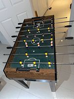 Full view of foosball table showing wood grain sides, green field, and players on metal rods with plastic handles