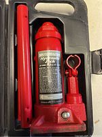 Red hydraulic bottle jack with pump handle and black plastic carrying case seen from top view.