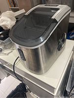 Side-angle view of stainless steel portable ice maker on white table with power cord attached.