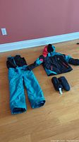 Full view of kids Spyder winter jacket, turquoise snow pants, and black mittens laid out on floor