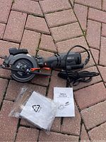 Photo of the Tacklife mini circular saw laid out on brick ground with power cord, blade visible, and instruction manuals alongside in plastic bag.