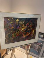 Frontal view of the abstract painting displaying vibrant swirling colors with white mat and acrylic frame, supported on an easel indoors.