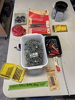 Top-down view of large plastic bin filled with assorted metallic screws, various packages of drill bits, holesaw, drywall bits and hardware containers on table.