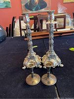 Pair of tall ornate silver plated candleholders seen from the front showing detailed designs and missing wax holders.