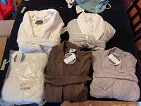 Top-down view of five folded terry cloth bathrobes in assorted colors laid on a table, showing tags and texture details.