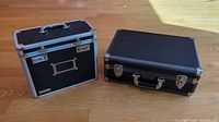 Two black professional hard cases displayed side by side on wood floor, Zeiko case with handle and lock hardware visible, Vaultz file box red background.