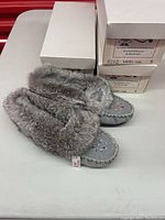 Three pairs of Bastein women's moccasins stacked in white labeled boxes next to one pair placed on table showing gray suede, fur trim and beading detail.