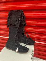 Side view of a pair of black suede moccasin boots showing double fringe and lace-up design on a white box in front of a red background.