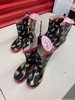 Three pairs of black rainboots with rainbow hearts pattern placed next to each other, all new with tags attached.