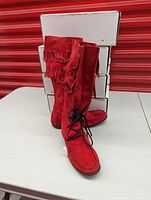 Front and side view of one pair of bright red women's moccasin boots with double fringe around the calf, black lace-up front, moccasin toe stitch, and rubber soles.