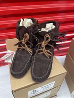 Front view of a pair of brown suede moccasin booties with double fringe and leather laces on top of a cardboard box.