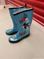 Pair of turquoise ladies rainboots with skull and heart with wings graphics and thick black rubber soles.