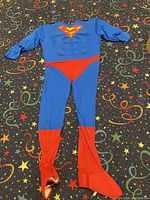 Blue Superman costume laid flat on star patterned carpet showing muscle padding and red boot cover attachments