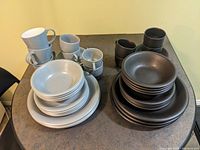 Photo showing two complete sets of Ikea stoneware dishes and cups in grey-blue and black colors arranged on a table.