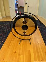 Front view of the Chinese brass gong on its curved black metal stand showing the painted Chinese characters and central black circle on the gong face.