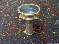 Overall side view of aluminum darabouka drum on carpet with colorful stars and spirals, showing its shape and reflective surface.