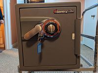 Front view of the gray SentrySafe with combination dial and key, showing the handle, lock, and brand logo.