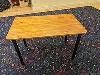 Full view of rectangular wooden table on colorful carpet, showing the tabletop with black legs and rounded edge corners designed to be flush with a wall.