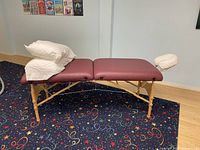 Full view of the lightweight massage table with burgundy padding, wooden frame, face cradle attached, and 2 white pillows on top.