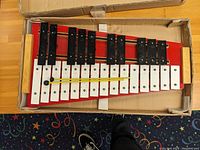 Top-down view of 25-note metal xylophone with two mallets, showing arrangement of white and black keys on red base inside cardboard box.
