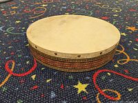 Middle Eastern frame drum lying flat on a patterned carpet, showing side view of wooden body decorated with geometric and stripe designs.