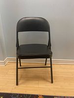 Front view of one black padded folding chair showing vinyl upholstered seat and backrest, metal frame, set on wooden floor.
