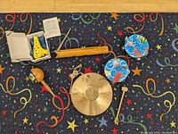 Top-down view of all items laid out showing ocarina in box, two tambourines, wooden guiro, maraca, brass gong and striker on patterned carpet
