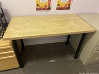 Rectangular MDF wooden table with light wood tone top and black metal legs, placed in a room with carpeted floor and white walls.