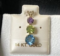Front view of the 14K gold pendant with three round-cut stones: amethyst (top), peridot (middle), blue topaz (bottom) displayed on white fabric backing with 14K gold marking.