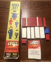 Photo of LEGO Doll Set 905 partial pieces arranged beside original box and instruction booklet on wood table. Box shows wear and tape on corners, bricks in red, white, and blue.