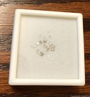 Loose assorted diamonds displayed in a white square box on a wooden surface, showing multiple small round diamonds.