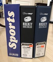 Side view of 3 collector albums, one labeled Sports and two Ultra Pro Hockey trading cards albums.