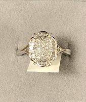 Ring displayed on gray padded surface showing top view of oval diamond cluster and surrounding baguette diamond halo.
