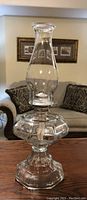 Photo showing vintage clear glass oil lantern measuring 18 inches tall on a couch background, in excellent condition.