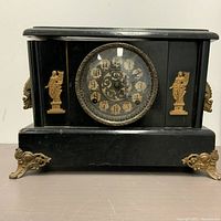 Front view of the black wooden antique mantel clock with brass feet and side handles, showing the round clock face with decorative bezel and aged dial.