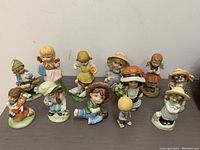 Front view of the twelve vintage bisque children figurines arranged on a table showing their attire and poses.