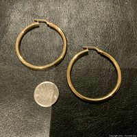 Both large 14K gold hoop earrings placed on a black leather surface next to a quarter for size reference.