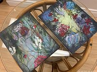 Pair of original oil paintings on canvas laid on a glass and wood table, showing abstract floral themes with vibrant colors, signed by the artist.