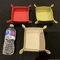 Three square-shaped handmade Italian leather valet trays in red, olive green, and beige on a black surface. A water bottle is placed to the left for size comparison.