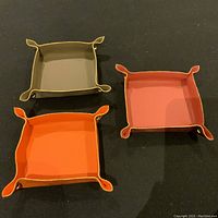 Three handmade Italian leather valet trays in gray, pink, and orange shades, arranged on black surface, showing shape, color, and gilded edges.