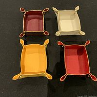 Overhead view of four handmade Italian leather valet trays in burgundy, cream, yellow, and red colors arranged in a square on a black background. Each tray has metal studded corners, visible leather texture, and slight variation in shape.