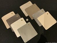 Eight handmade leather coasters arranged in two rows on black fabric. Each coaster is square with metal eyelet in top right corner, colors range from cream beige to dark brown and gray.