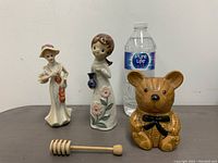 Three vintage figurines with two ceramic girl figures and one teddy bear figure placed next to a water bottle for scale, including a wooden honey dipper.